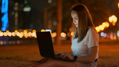 A-young-female-student-with-a-laptop-at-night-in-the-city-looking-at-the-computer-screen-and-typing-with-his-hands-on-the-keyboard.-Remote-work-on-the-Internet.-The-student-does-the-work.-Night-life-in-the-city